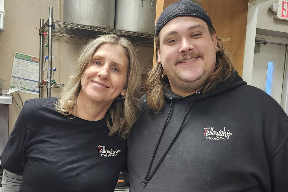 Two people in Fellowship Missions shirts stand together in a kitchen.