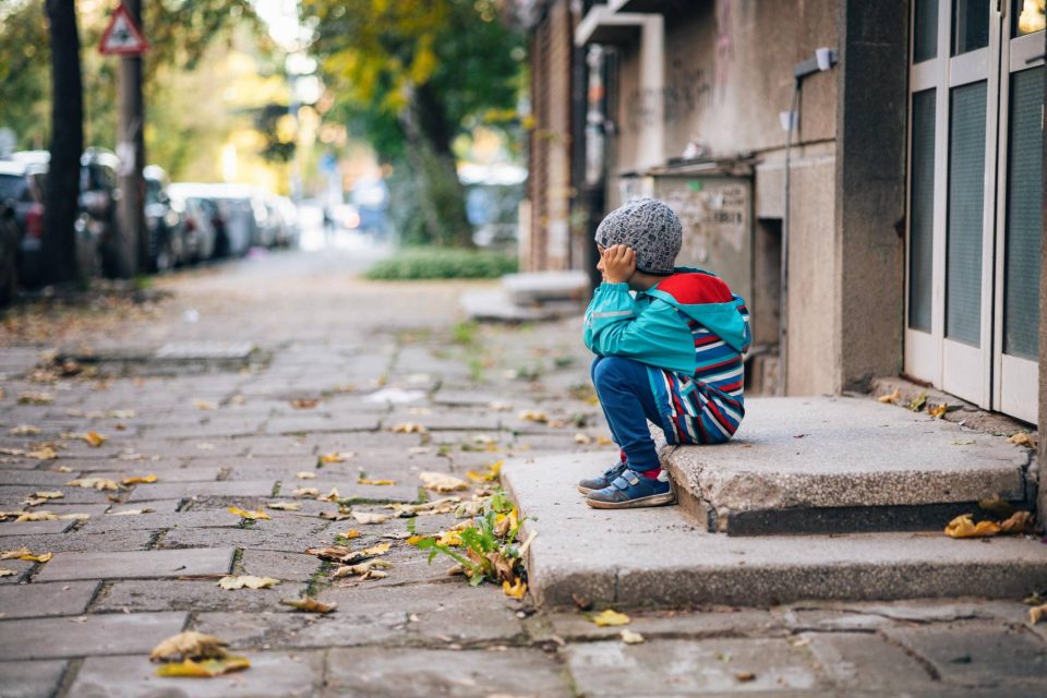 A homeless child in a striped jacket sits alone on a city sidewalk doorstep.