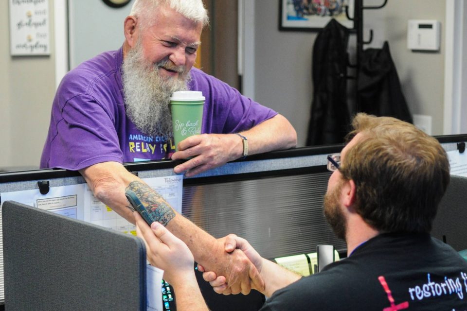 Two men shaking hands across an office cubicle, both smiling and friendly.