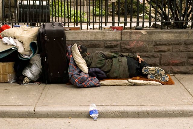 Unhoused person sleeping on sidewalk, suitcase and blankets nearby.
