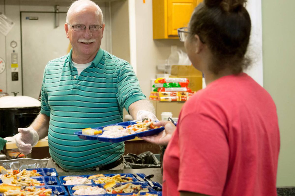 alt=Caucasian man smiling as he serves food to a woman in a pink shirt at the emergency homeless shelter